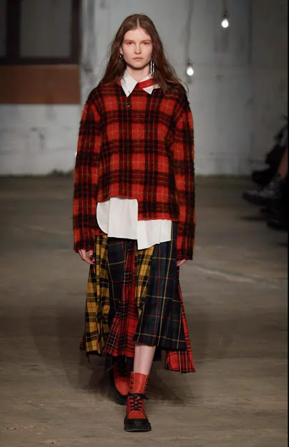 The woman wears a sweater and plaid long skirt.