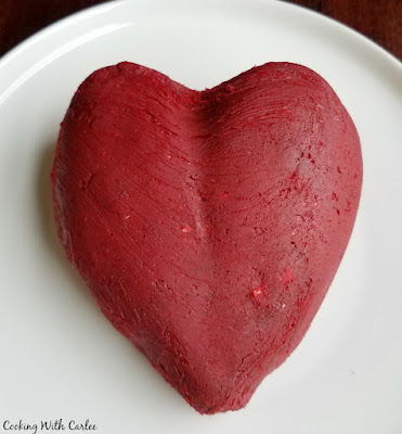 Luscious red velvet cake and cream cheese frosting flavors come together for a one of a kind dessert experience. Red velvet cheese balls are perfect for Valentine's Day, Christmas or just because! red velvet cheese ball hand shaped into a heart shape, ready to be coated in white chocolate