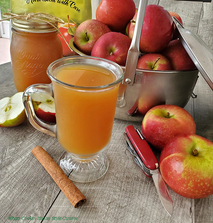 this is a photo of homemade apple-cider in a glass cup with apples in a big pot, mason jar filled with cider and sliced and quartered apples in the photo with a knife to slice them
