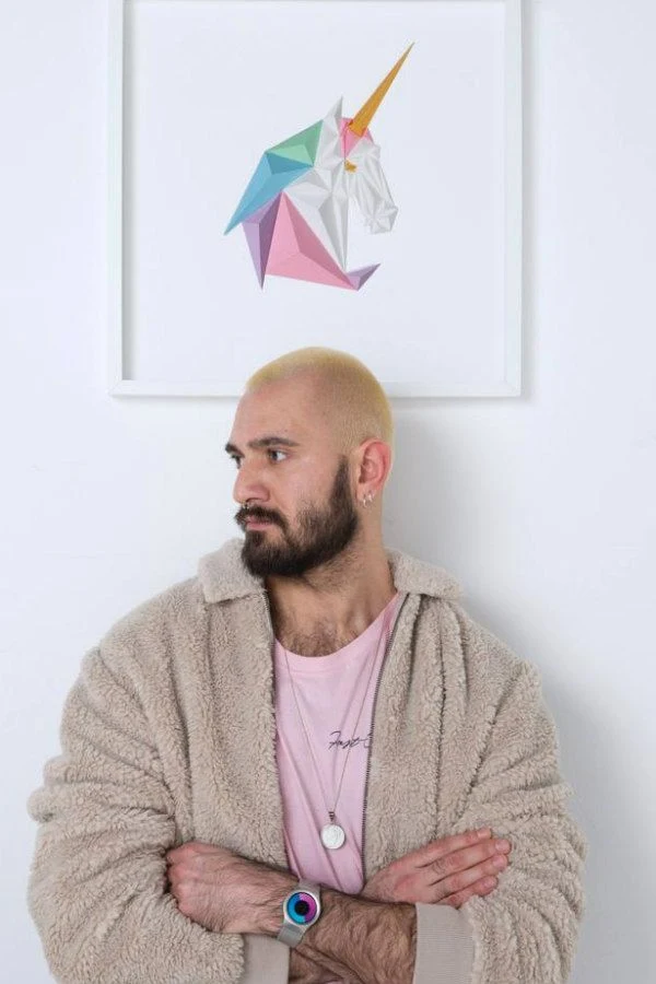 artist Tayfun Tinmaz standing in front of a pastel 3D folded paper unicorn in square white frame hanging on wall