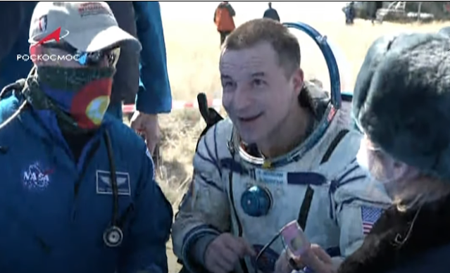 A screen capture of Andrew Morgan back on Earth being examined by a Dr. Scheuring, who is wearing a bandana around his face and a khaki ball cap.