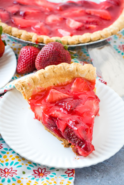 slice of strawberry pie made with fresh strawberries and gelatin with a whole pie in the background with fresh strawberries around on a colorful napkin