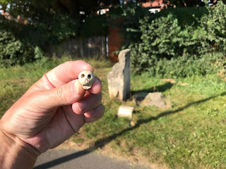 A picture showing a hand holding a small, ceramic skull, Skulferatu #50, with the Hob Stone and Plague Stone in the background.  Photo by Kevin Nosferatu for the Skulferatu Project.