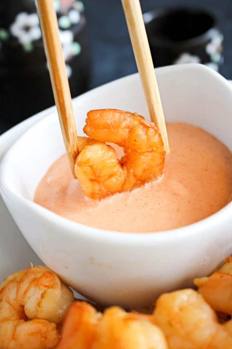 Side view of chopsticks holding a shrimp being dipped in yum yum sauce in a white bowl.