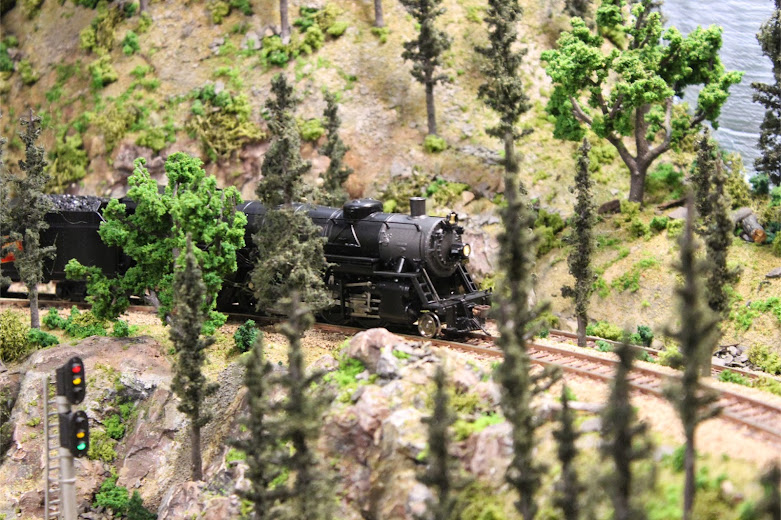 A USRA Light Mikado 2-8-2 steam locomotive passing through a mountain forest scene with rock outcroppings and trees