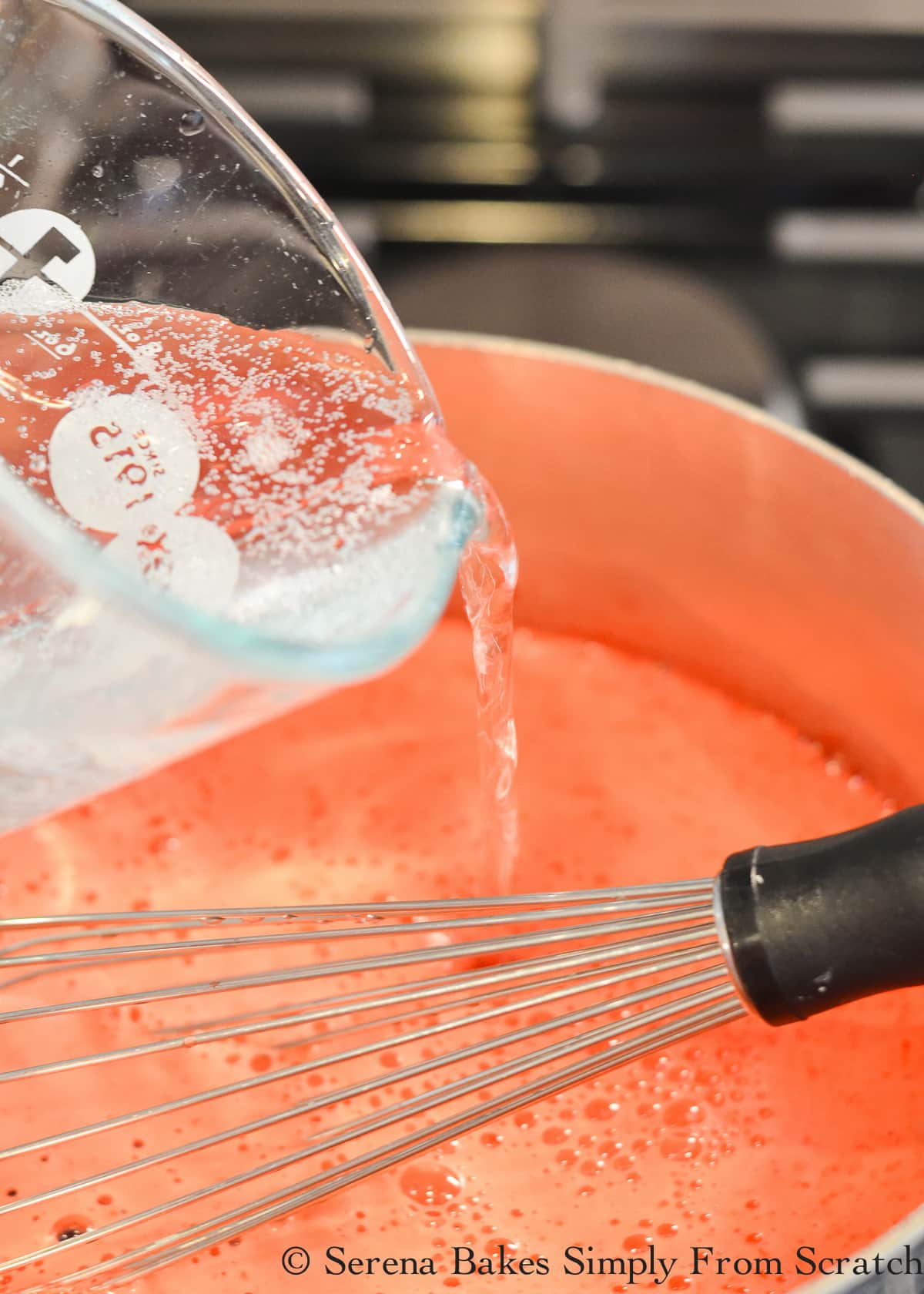 7 Up in a measuring glass being poured into Raspberry Jello mixture in a stainless steel pot.