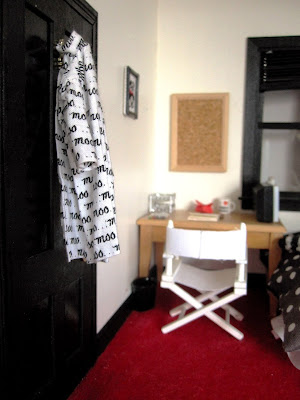 1/12 scale modern miniature bedroom scene with a black and white 'moo' dressing gown hanging from a black wardrobe door . In the background is a white director's chair at a desk, with a portable TV on the desk pointing towards the bed.