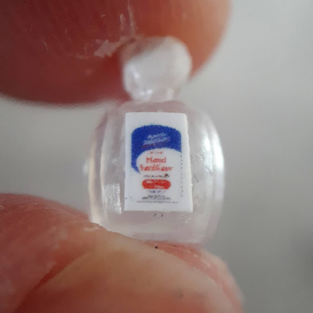 Tiny one-twelfth scale bottel of hand sanitiser held between a very large finger and thumb.