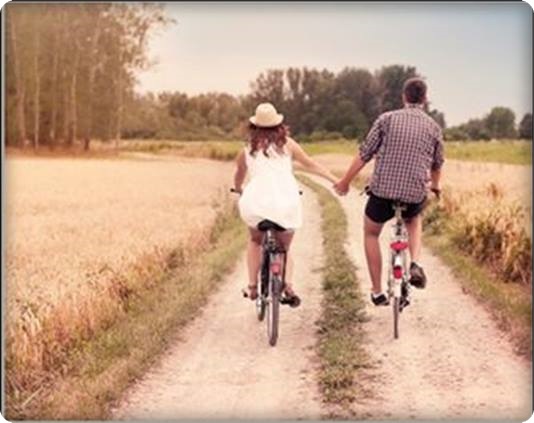 Casal pedalando de bicicletas de mãos dadas entre os campos.
