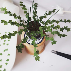 1/12 scale miniature plant kit with half bare and half leafy stems in a plant pot filled with 'soil' on top of a cotton reel, with tray of extral leaves on one side and a pair of pliers on the other.