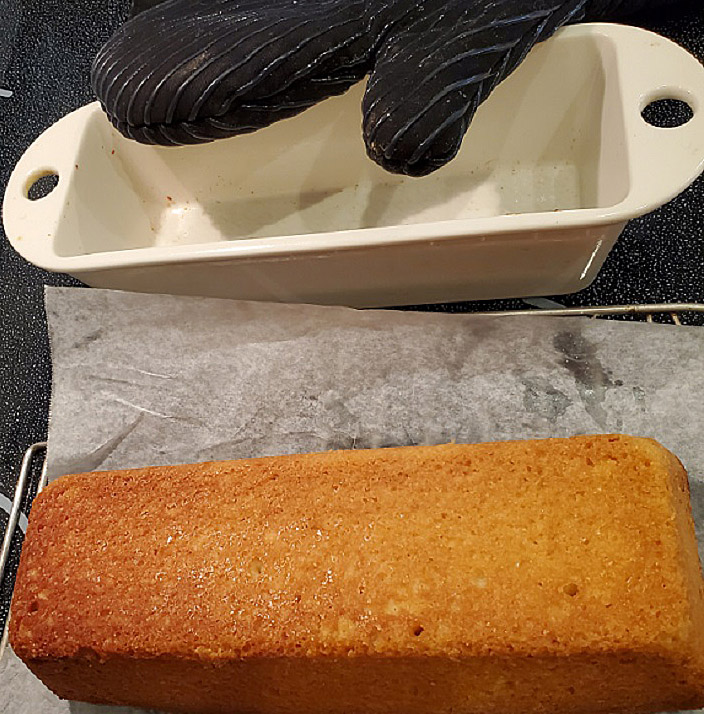This is a lemon loaf cake on wax paper and a wire rack cooling with a black pot holder and the empty Revol porcelain pan it was cooked in