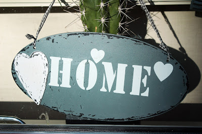 ID: an oval sign that reads in white text on a light green background "Home" it is surrounded by hearts. The sign is hanging from a green cactus.