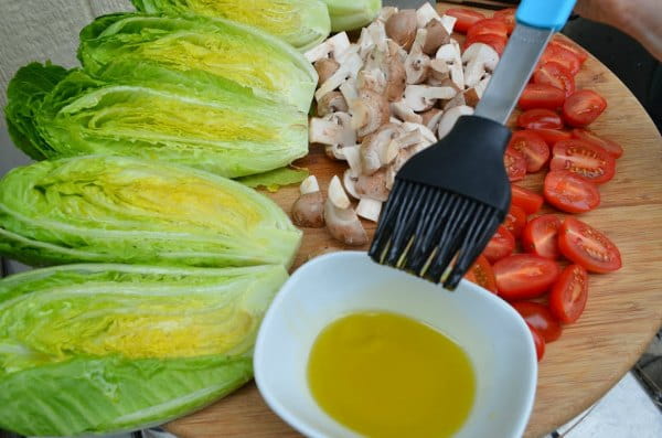 Halved Romaine Hearts being brushed with olive oil with mushrooms and tomatoes on a tray for Grilled Romaine Salad.