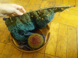 A lace shawl held live on the needles.  The shawl is knit in a gradient from purple to yellow-green.  Both the yarn cake and the end of the shawl are tucked into a wooden yarn bowl.  The shawl has a number of stitch markers looped around the needle.