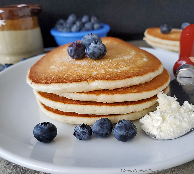 this is a photo of buttermilk pancakes stacked up with blueberries on top. There is a wooden handled butter knife on the white plate with a pitcher of syrup and more pancakes on a plate in the background along with a bowl of blueberries in a small white ceramic bowl