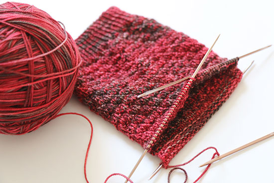 Hand knit red wool socks in progress on metal double point knitting needles next to a ball of yarn on a white background.