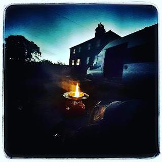 Dusk sky, outline of house in background, foreground is a campfire