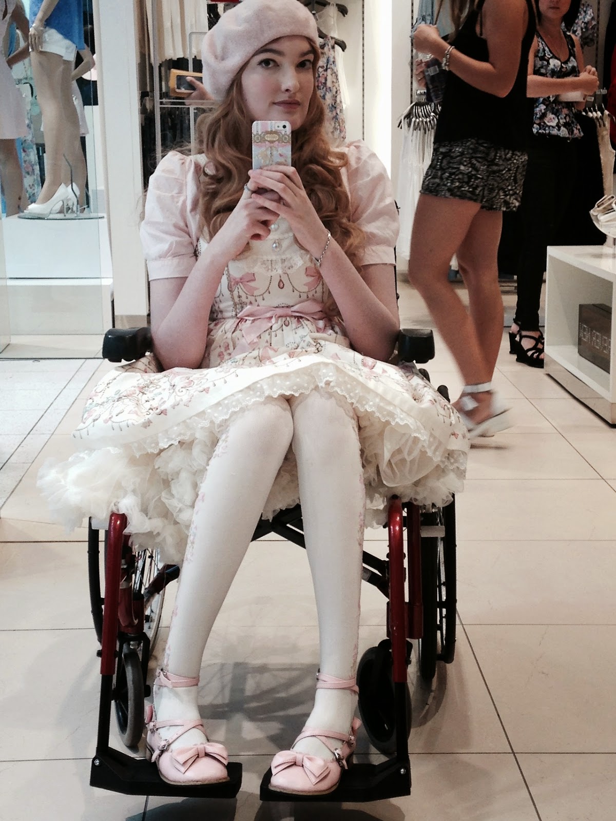 Young white woman sitting in a wheelchair taking a selfie with a smartphone, wearing a white frock dress and light pink hat