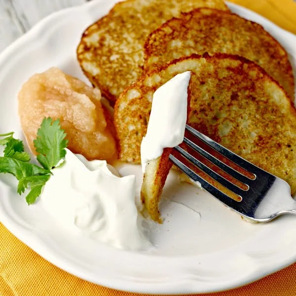 A piece of Easy Potato Pancakes by Renee's Kitchen Adventures on a fork with sour cream. Applesauce and sour cream on plate with pancakes in background. 