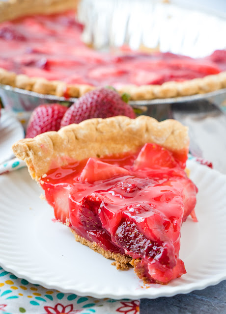 Slice of fresh strawberry pie on white plate with whole strawberry pie in the background with fresh strawberries around the pie and a colorful napkin