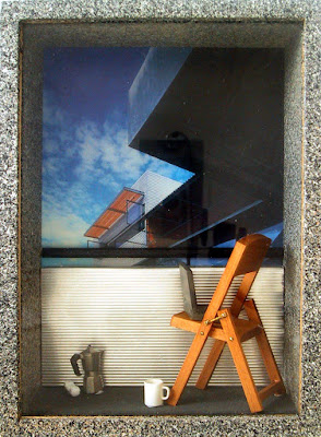 A one-twelfth scale modern miniature city balcony scene in a box frame with sprayed concrete walls and floor and a corrugated iron balcony wall. On the balcony is a folding wooden chair with a laptop on it. On the floor is a cordless phone, a mug and a stovetop espresso jug.