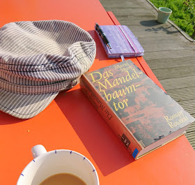 Buch "Das Mandelbaumtor" auf orangem Gartentisch mit Kaffee, Gartenmütze und Notizbuch