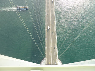 Looking down and the road deck from the top of one of the main towers on the Akashi Kaikyo bridge, There are cables supporting the bridge visible as well as a boat in the sea about to cross underneath the bridge