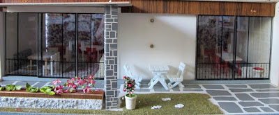 Vintage dolls' house patio with stonework pillars and paving, and planter box full of plants. On the paving is a set of white outdoor table and chairs.