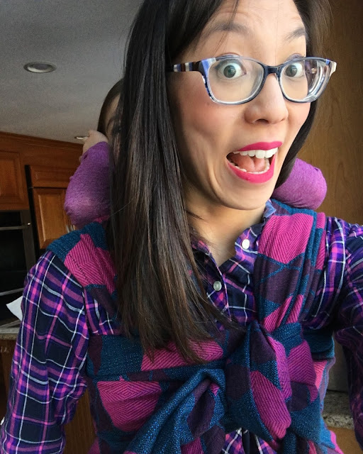 [Image of a tan skin bespectacled Asian woman wearing a disgruntled child on her back in a pink and blue largescale argyle patterned woven wrap carrier. Woman has excited eyebrows and bright pink lips in an open smile that says “no problem, I’ve got this.” Woman’s has on a purple, navy, and pink plaid top.]