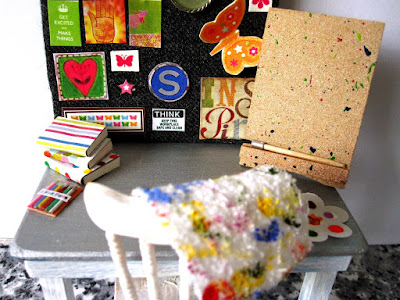 Studio chair and table with an easel, paints and cleaning cloth, a pile of noteboks next to some pencils and a noticeboard pinned with inspiration.