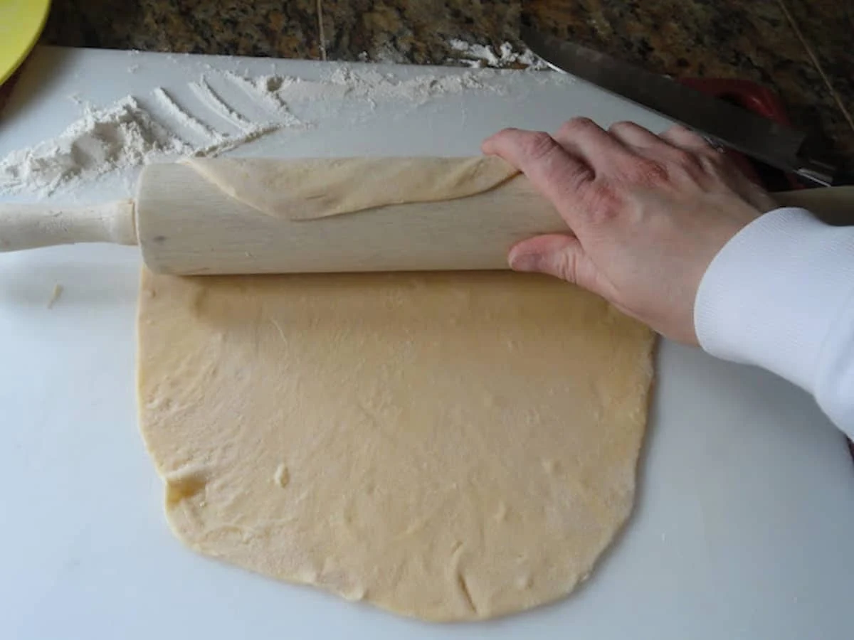 Homemade Egg Noodle dough being rolled out with a rolling pin on a white cutting board.