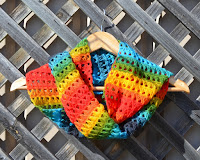 Cowl displayed on wooden coathanger as if it were around the neck. The cowl has one twist in it and two layers around the neck. Rainbow stripes are in the following sequence dark blue, light blue, green yellow, orange, red, and then in reverse.  The background is a wooden trellis of diagonal slats.