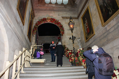 Photo of a large staircase in a country house. It is decorated for Christmas. Along the left side of the stairs, is a platform stairlift, carrying a wheelchair user up the stairs.