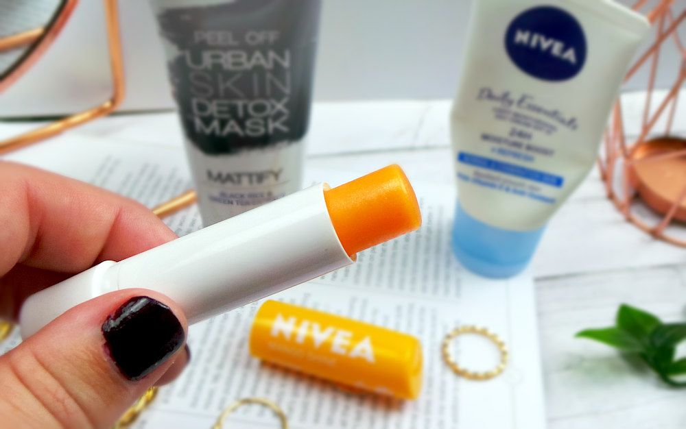 Image shows my hand holding a white tube with an orange lip balm in it. The background is out of focus with a mirror, two tubes of skincare, a magazine and rings