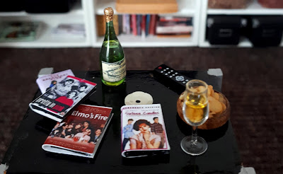 One twelfth scale modern miniature lounge scene with a coffee table in the foreground holding several DVDs of films from the 1980s (including Sixteen candles, with the DVD out of the cover, Pretty in pink and the Breakfast club), a TV remote control, a glass and bottle of sparkling wine and  a bowl of potato chips.