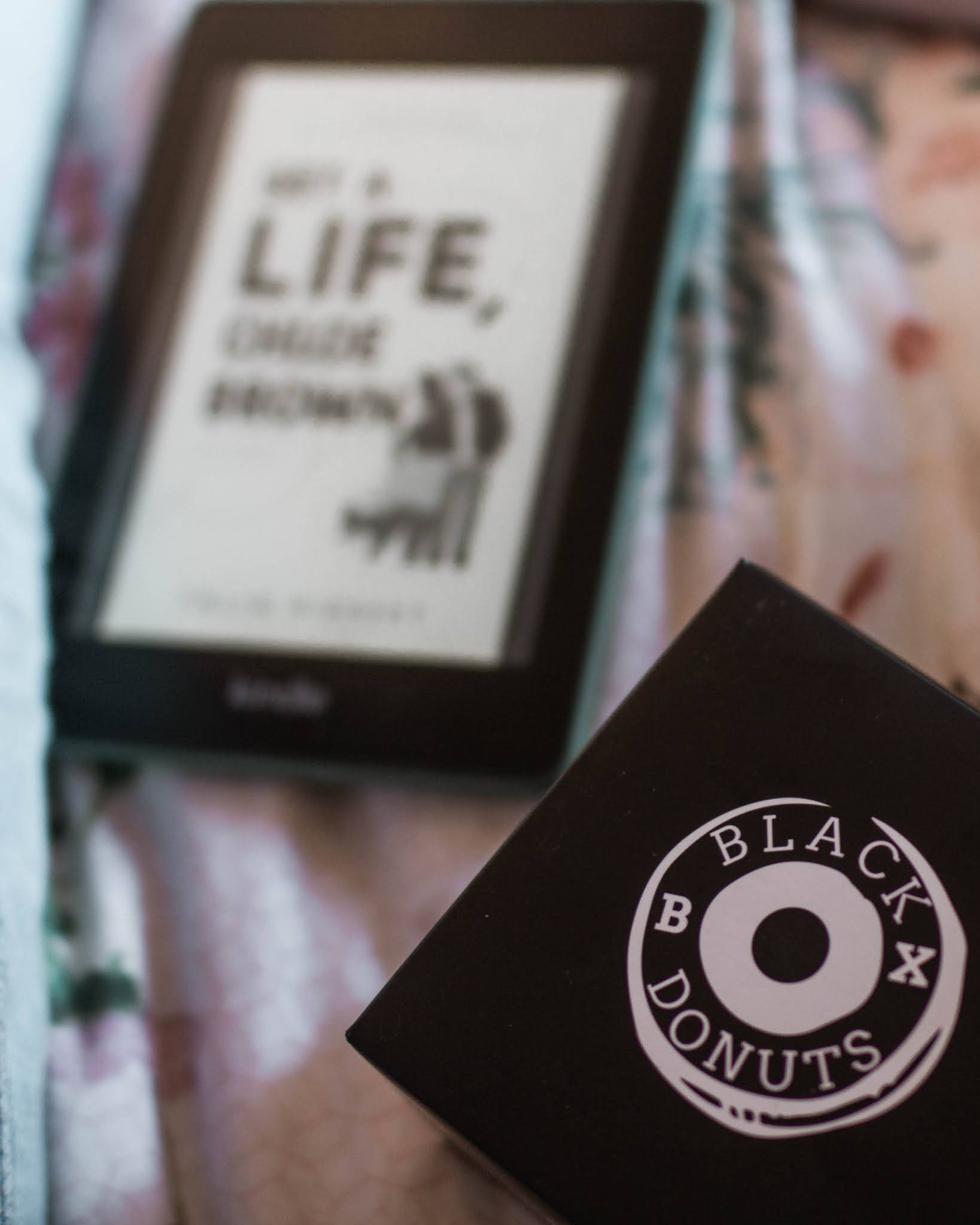 kindle displaying get a life chloe brown by talia hibbert cover on screen and a donut box sitting on pink bedding