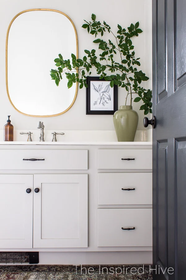 Master bathroom with greige vanity cabinets, oil rubbed bronze hardware, nickel faucet, and brass mirror. Large green vase with oversized greenery.