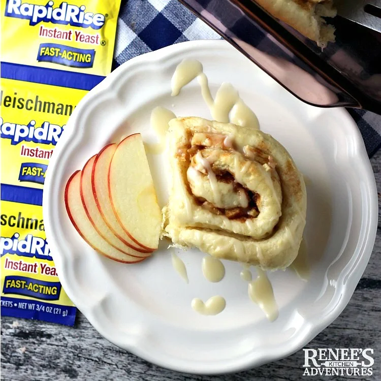 Apple Cinnamon Rolls with Maple Glaze by Renee's Kitchen Adventures on a white plate with apples in the background and package of yeast next to roll.