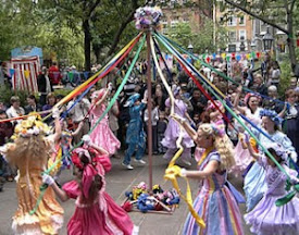 Girls in multi-colored dresses dance around a Maypole at a festival.
