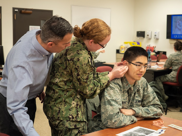 Three people, one seated at a table (far right). The man on the left supervises the woman in the middle, who is placing an acupuncture needle on the seated man.