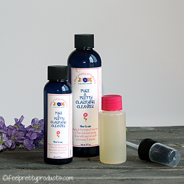 Three different sizes of Pure & Pretty Clarifying Cleanser sitting on a wooden table with purple flowers behind them