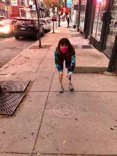 Me crouching down with a mask on, in front of a chalked happy face on the sidewalk, also wearing a mask.