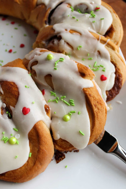 Soft and sweet cinnamon rolls take on a festive wreath shape. They are a perfect holiday breakfast or something fabulous for overnight guests. close up of knife removing a cinnamon roll from the wreath
