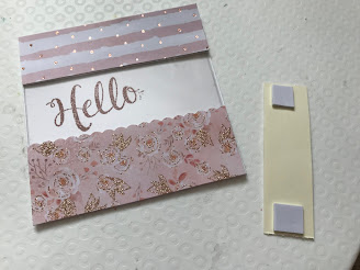 Front of a square card. In the middle is a cut out section with acetate and you can see the stamped 'hello'. Above the acetate it is now decorated with pink and white patterned paper with rose gold dots. Below the acetate is a pink and rose gold glitter floral patterned paper. Next to this is a thin rectangle of cream card which has 3D sticky pads stuck to the top and bottom.