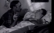 A thin, middle-aged white man sits at the bedside of a disabled young woman, looking into her face and holding her hand