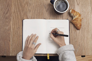 Journal notebook on a wooden table with hands writing, coffee and croissant on the side