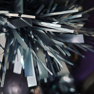 Petrol-grey tinsel Grey tinsel above a blurred purple bauble and against the background of a dark purple wall. The fronds of the tinsel are quite chunky, the tinsel is dark grey and metallic