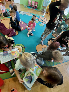 Przedszkolaki siedzą na dywanie, czytają i oglądają książki o Kubusiu Puchatku. Tło: kolorowa sala przedszkolna.