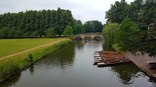 On the left of the photo you can see a large expanse of green space which borders the river Cam which is meandering through the centre of the picture. In the distance you can see the triple arches of Trinity Bridge spanning the river. On the right of the picture you can see 10 punts moored.