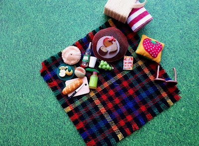 Aerial view of a one twelfth scale picnic on the grass with a picnic rug, basket with a towel, various items of food, a bottle of wine and a book and a cushion.